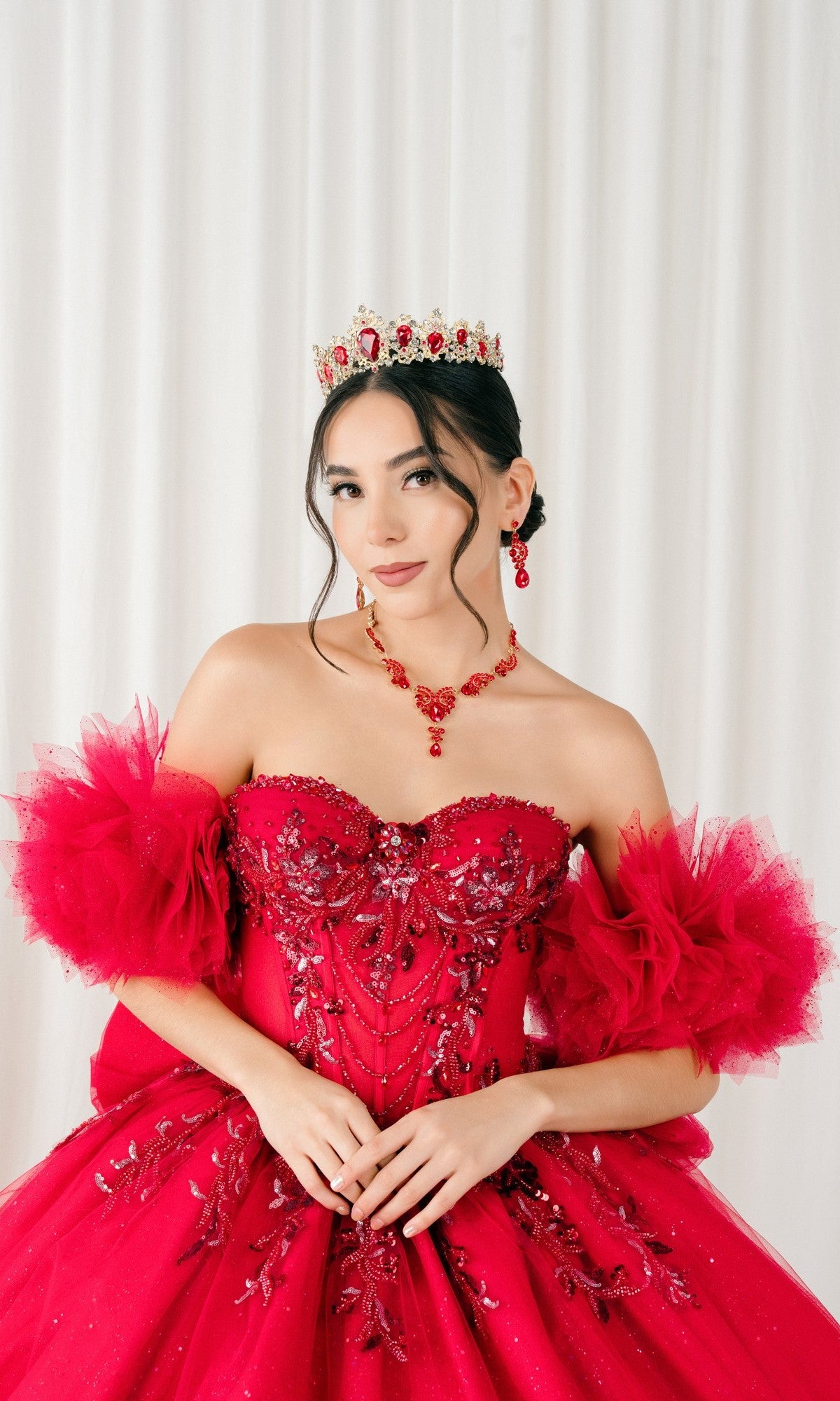 A woman wearing the Quinceanera Dress 8380J By Cinderella Couture, a bright red ball gown with a corset bodice and embellished details, stands in front of white curtains, accessorized with a matching crown and jewelry.